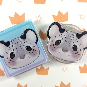 May include: Two clear acrylic pins featuring a cartoon snow leopard face. One pin is square with a light blue background, the other is round. The snow leopard has black ears, spots, and a pink nose.