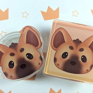 Könnte beinhalten: Zwei transparente Acrylstifte mit einem Cartoon-Hyänen-Gesicht mit braunem und hellbraunem Fell, schwarzen Augen und dunklen Flecken. Ein Stift ist rund, der andere quadratisch. Der Hintergrund ist weiß mit orangefarbenen Kronen- und Sternillustrationen.