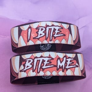 Puede incluir: Dos pulseras de tela con un diseño de boca de dibujos animados. Una pulsera dice "I BITE" y la otra "BITE ME" en una fuente audaz y estilizada. Las pulseras presentan una combinación de colores rojo y blanco con un logotipo negro.