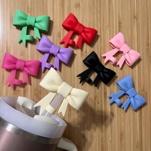 May include: Assortment of silicone bow-shaped straw covers in various colors, including green, red, purple, pink, white, black, and blue. The covers are designed to fit over a straw and add a decorative touch to a beverage.