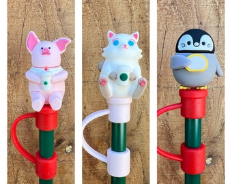 May include: Three different colorful bottle stoppers with a pink pig, a white cat, and a gray penguin. Each stopper has a green stem and a red base.