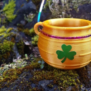 Könnte beinhalten: Ein goldener Topf mit einem grünen Kleeblatt-Design, der an einer regenbogenfarbenen Schnur hängt. Der Topf steht auf einem Moosbett.