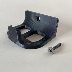 Puede incluir: Un soporte de pared de plástico negro con un agujero para tornillo y un diseño curvo. Está diseñado para sujetar un objeto pequeño, como un teléfono o una tableta. Se incluye un tornillo negro.