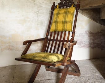 May include: A vintage wooden folding chair with a yellow and green plaid cushion and backrest. The chair has a carved top and armrests. The chair is positioned under a wooden staircase in a room with stone flooring and aged walls.