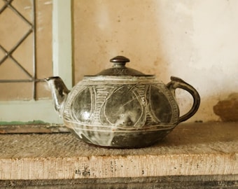 May include: A ceramic teapot with a dark gray glaze and a decorative pattern of stylized leaves. The teapot has a curved handle and spout, and a matching lid. It sits on a weathered wooden surface near a window.