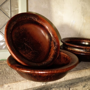 Puede incluir: Una pila de tres cuencos marrones de aspecto rústico con un acabado brillante. Los cuencos están hechos de un material marrón oscuro, posiblemente cerámica o madera, y tienen un diseño simple y redondeado. Los cuencos están apilados.