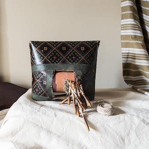 May include: A dark, patterned textile object with a rectangular opening, possibly a case or container. The object is decorated with a repeating geometric design in shades of black, green, and red. A collection of wooden tools and a spool of thread are in front of the object.