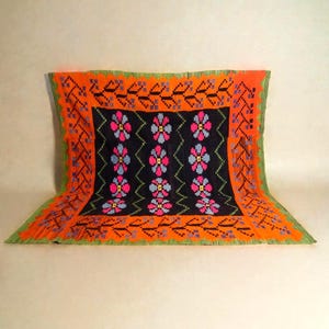 May include: A colorful, hand-stitched textile with a black central field featuring rows of stylized flowers in pink, blue, and yellow. The border is orange with green trim. The textile is folded, displaying its intricate design and craftsmanship.