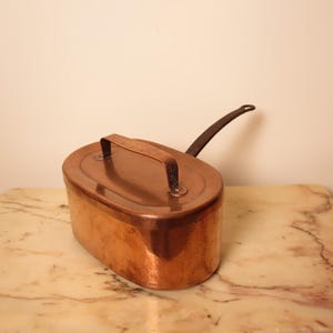 May include: A copper oval-shaped cooking pot with a lid and a long handle. The pot has a rectangular handle on the lid. The pot is sitting on a marble-patterned surface.