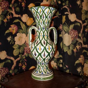 Könnte beinhalten: Eine weiße Keramikvase mit grünen, braunen und gelben geometrischen Mustern. Die Vase hat zwei Henkel und eine breite Öffnung.