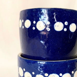 May include: Two blue ceramic mugs with white polka dots stacked on top of each other. The mugs are a deep blue color and have a glossy finish.