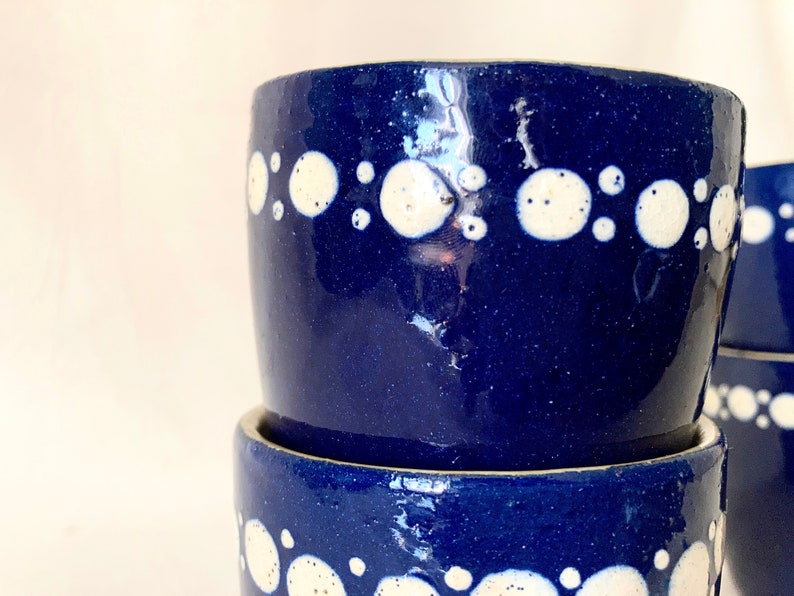 May include: Two blue ceramic mugs with white polka dots stacked on top of each other. The mugs are a deep blue color and have a glossy finish.
