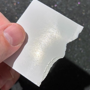 May include: A small, square piece of white paper with a smooth, reflective, pearlescent finish. One edge of the paper is straight, while the opposite edge features a rough, torn, and uneven texture, revealing the fibrous layers of the material.