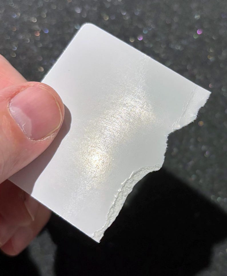May include: A small, square piece of white paper with a smooth, reflective, pearlescent finish. One edge of the paper is straight, while the opposite edge features a rough, torn, and uneven texture, revealing the fibrous layers of the material.