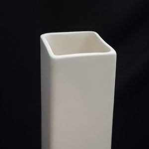 May include: A white ceramic vase with a square shape and a slightly flared top. The vase has a smooth, matte finish and sits on four small feet.