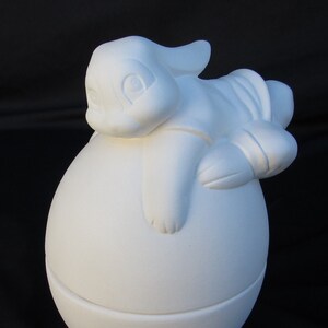 May include: White ceramic Easter egg with a bunny sitting on top. The bunny has a long, floppy ear and is looking to the side. The egg is decorated with a grass-like base.