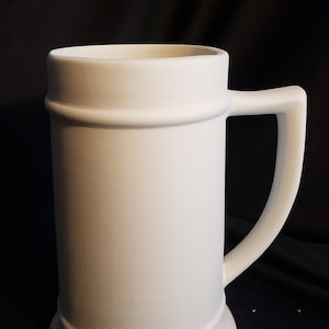 May include: White ceramic beer stein with a handle. The stein has a wide base and a cylindrical body with a raised lip around the top.