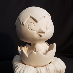May include: White ceramic figurine of a baby chick hatching from an egg, sitting in a nest. The chick has large eyes and a beak.