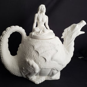 May include: White ceramic teapot shaped like a dragon with a woman figurine on top of the lid. The teapot has a detailed dragon design and the woman is wearing a flowing dress.