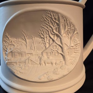 May include: A cream-colored ceramic teapot with a lid and handle. The teapot features a circular bas-relief scene of a winter landscape with a house, trees, and figures. The design is detailed and adds a decorative element to the teapot.