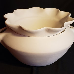 May include: A white ceramic serving bowl with a ruffled edge lid. The bowl has two handles and a smooth, matte finish. The lid has a scalloped edge. The bowl is designed for serving food.