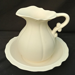 May include: A white ceramic pitcher with a fluted design and a decorative handle sits on a matching white ceramic saucer.