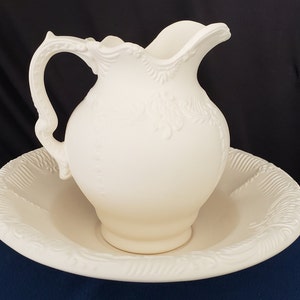 May include: White ceramic pitcher and matching bowl with intricate embossed designs. The pitcher has a handle and a wide mouth. The bowl is round and has a raised rim.