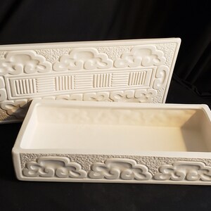 May include: White ceramic box with a lid. The box has a raised design of clouds and geometric patterns. The lid is open and the inside of the box is empty.