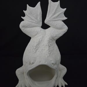 May include: A white ceramic frog sculpture with its mouth open and arms raised. The frog has textured skin and webbed feet. The sculpture is set against a black background.