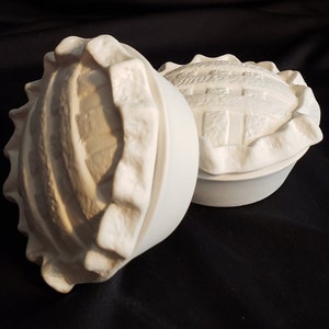 May include: Two white ceramic pie crusts, one with a raised, textured design and the other with a smooth surface. Both are in the shape of a pie crust.
