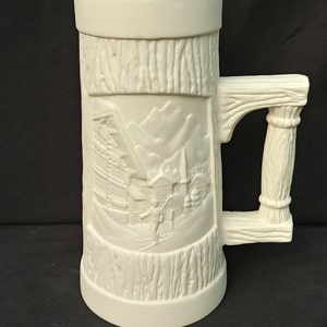 May include: White ceramic beer stein with a raised design of a winter scene with a skier and a chalet. The stein has a wooden-style handle.