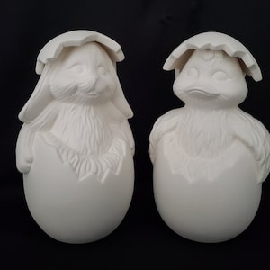 May include: Two ceramic figurines of animals hatching from eggs. The figurine on the left is a rabbit, and the figurine on the right is a chick. Both figurines are white and have a textured surface.
