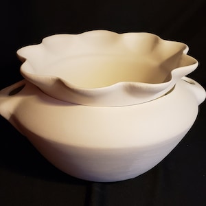 May include: A two-piece, off-white ceramic serving bowl set. The bottom bowl has two handles and a rounded shape. The top bowl has a scalloped edge and sits inside the bottom bowl. The set is unpainted and ready for customization.