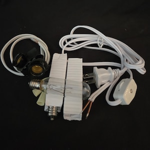 May include: A collection of electrical components, including two black light bulb sockets, two clear light bulbs, a white power cord with a plug, and a white inline switch. The components are arranged on a black surface.
