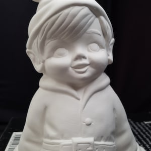 May include: White ceramic figurine of a sitting elf wearing a hat with a bell on top. The elf has a smiling face and is wearing a jacket with a belt.