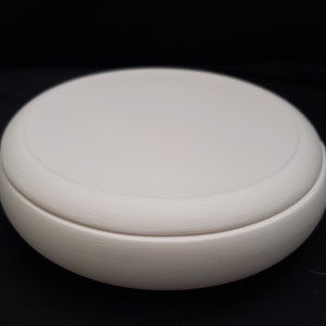 May include: A round, off-white ceramic box with a lid. The box has a smooth, matte finish and a simple, minimalist design. The lid fits snugly on top of the base, creating a seamless look. The box is set against a black background.