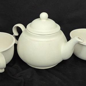 May include: A white ceramic teapot with a lid and two matching cups. The teapot has a round body and a spout. The cups have a rounded shape and a handle.