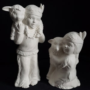 May include: Two unpainted ceramic figurines. One depicts a child wearing a feathered headdress and carrying a lamb on their shoulders. The other figurine shows a child with a lamb. Both are in a Native American theme.