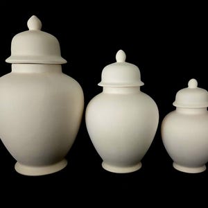 May include: Three off-white ceramic ginger jars with matching lids. The jars vary in size, with a rounded body and a small knob on top of each lid. The jars are displayed against a black background.