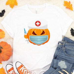 May include: White t-shirt with a pumpkin wearing a nurse's hat and a face mask. The pumpkin is holding a syringe. The shirt is laying on a white surface with a pair of orange converse shoes, a pair of blue jeans, a fake skull, and fall leaves.