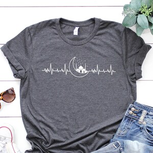May include: A dark grey t-shirt featuring a white crescent moon and mosque design, with a heartbeat line on either side of the design. The shirt is laid flat on a white wooden surface with a pair of sunglasses, white trainers, and blue jeans.