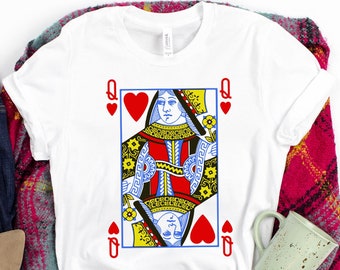 May include: White t-shirt featuring a graphic of the Queen of Hearts playing card. The shirt is folded and displayed on a pink and white checked blanket with a pair of brown sandals and blue jeans.