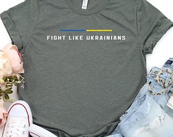 May include: A green t-shirt with the text "Fight Like Ukrainians" in white letters. The shirt has a blue and yellow stripe across the chest, representing the Ukrainian flag. The shirt is laid out on a white surface with a pair of white sneakers and blue jeans.