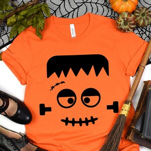 May include: Orange t-shirt with a black Frankenstein face graphic. The shirt is laying flat on a white background with a broom, books, and a black spiderweb.