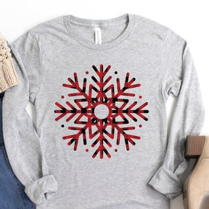 May include: A gray long-sleeve shirt with a red and black plaid snowflake design. The shirt is paired with blue jeans and brown ankle boots.