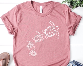 May include: A dusty rose t-shirt with a white outline of four sea turtles swimming in a line. The shirt is laying flat on a white wooden surface with a pair of brown sandals and blue jeans.