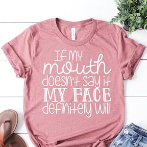 May include: A dusty rose t-shirt with the text "IF MY mouth doesn't say it MY FACE definitely WILL" in white script. The shirt is paired with blue jeans and brown sandals. A small potted plant is also in the image.