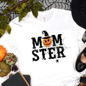 May include: White t-shirt with a black and orange pumpkin graphic and the text "MOMSTER" in black. The shirt is surrounded by Halloween decorations, including a witch's hat, black shoes, and orange and green pumpkins.