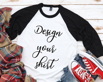 Puede incluir: Una camiseta de béisbol blanca y negra con el texto "Design your shirt" en escritura negra. La camiseta está sobre un fondo de madera blanca con una bufanda de cuadros rojos y negros, zapatillas rojas y blancas y unos vaqueros azules.
