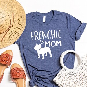 May include: A navy blue t-shirt with the text "Frenchie Mom" and a white silhouette of a French Bulldog. The shirt is laid out on a white surface with a straw hat, brown sandals, and a white crochet bag.